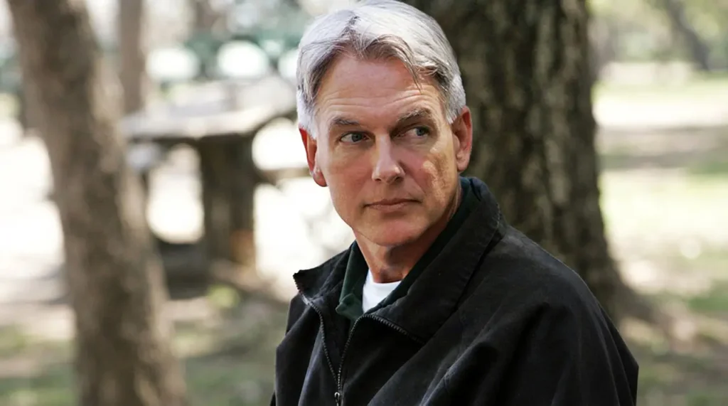 A medium shot photograph of actor Mark Harmon in character as Leroy Jethro Gibbs from NCIS, wearing his signature black jacket and green polo shirt, looking thoughtfully off-camera with a neutral expression.