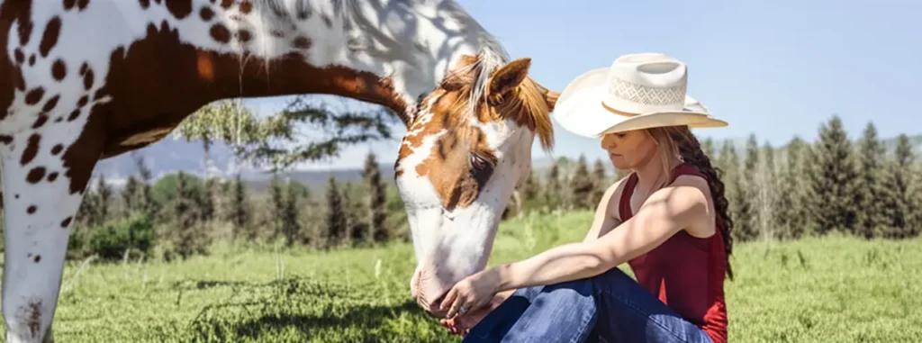 Actress Amber Marshall wearing a cowboy hat sits in a grassy field gently touching the nose of a brown and white paint horse on the set of the television show Heartland.