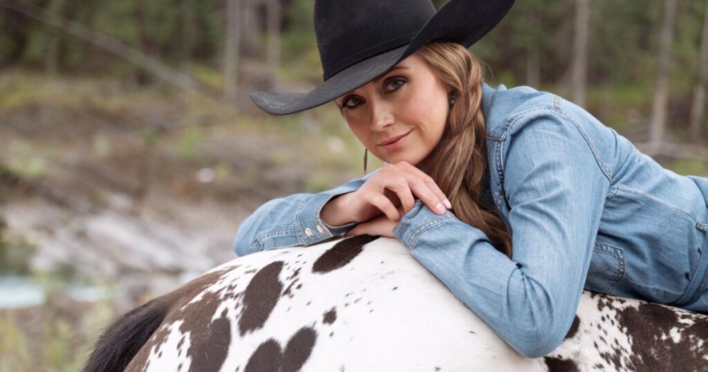 Amber Marshall wearing a cowboy hat is lying on the back of a spotted horse.